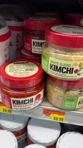 The video shows a close up view of various jars of kimchi on grocery store shelves. The jars have red lids and are labeled “Kimchi” in white lettering with a red background. Some of the jars are labeled as "Sliced Kimchi" and "White Sliced Kimchi." The bottom shelf contains larger jars labeled "Kimchi Sliced Cabbage." A price tag is visible: $6.99. There are some other food products on the shelves around the kimchi jars.
