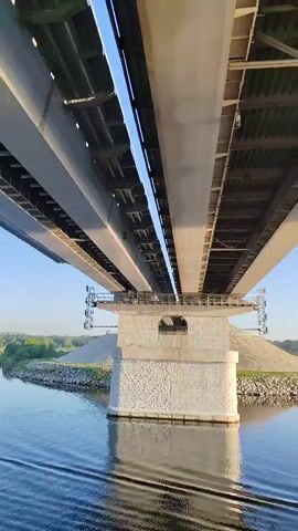 The camera, low to the water, moves underneath a massive gray bridge. The bridge's structure is visible, featuring beams, supports, and rectangular openings in the central pillar. The supporting pillar is made of white stone. The surrounding environment includes a body of water with gentle ripples, a rocky shoreline with some green vegetation, and a large mound of earth visible in the background. The sky above is a clear, bright blue.