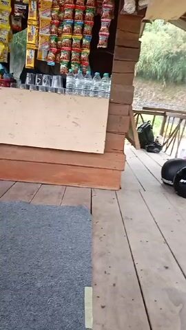 The camera pans from left to right in what appears to be an outdoor rest stop. There is a snack stand selling chips and drinks in the beginning, with some glasses lined up on the countertop. A wooden plank floor runs through the scene. Two people sit on the right of the frame, facing each other. There are three helmets and various bags and personal items scattered around.  Outside the rest stop are three motorbikes parked on a dirt lot next to a road, against a green, sloped landscape in the background.