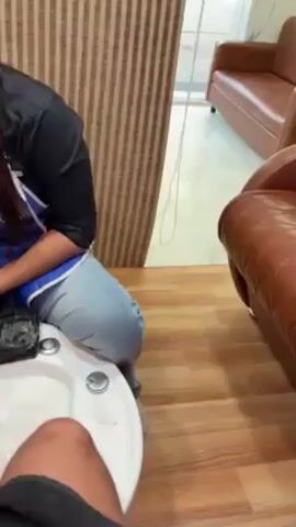 A woman in a blue apron with the words "Caprabo Brazilian Keratin" embroidered on it is giving a pedicure to someone. She is using clippers and a cuticle pusher to work on the toenails. The person receiving the pedicure is sitting in a chair with their foot in a small white tub of water. The room has wood flooring and a striped wall covering. There is a brown leather couch in the background.
