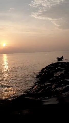 The video shows a serene seaside scene at sunset, with the sun low on the horizon, reflecting on the calm water. In the foreground, a rock breakwater extends into the water, composed of large, dark stones. A small fishing boat with a few figures aboard can be seen floating some distance away, adding to the tranquil atmosphere. A second, even smaller boat is further in the distance. The sky is a mix of pale orange and muted gray hues, suggesting the sun is setting.