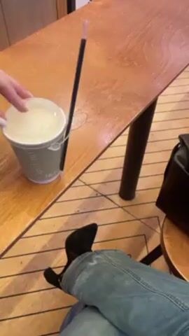 Here's a description of the video:
A person sits at a wooden table with a drink in a light green paper cup with a lid and a straw. The person is wearing blue jeans and black suede boots. Their hands are visible; they have white nail polish. The drink is sitting on the table next to a black purse. In the background, there is a blurry view of a coffee machine and other equipment. The floor has a pattern of dark and light wood. The person moves the cup on the table.