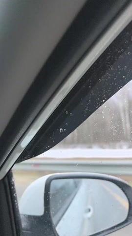 The video is shot from inside a car on a rainy day. The view out the passenger side window shows raindrops on the plastic window visor and on the window itself. The side view mirror is visible, showing the road behind the car. Snow is visible on the side of the road. The sounds of music can be heard.
