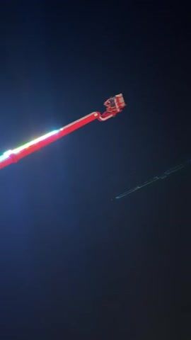 The video displays a thrilling amusement park ride at night. A large red and white mechanical arm with attached gondolas spins and swings visitors high into the air against a dark sky. Bright LED lights illuminate the ride, creating a vibrant spectacle. In the background, buildings and a large sign reading "WELCOME TO SUNNY BEACH" are visible. The camera captures the dynamic movements of the ride, emphasizing the sense of excitement and exhilaration.