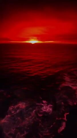 This is a vertical video of a crimson ocean under a red sky with a setting sun. The sun appears as a bright, circular orange orb on the horizon. The water is a deep red, with darker patches and foamy, lighter red waves crashing in the foreground. The overall effect is one of intense, fiery color. There are no people or man-made objects visible. The scene is solely comprised of the ocean and sky during what appears to be a dramatic sunset or sunrise.