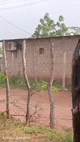 The video presents a static, medium shot of a rural scene. In the foreground, a rustic fence is constructed from roughly hewn wooden posts. Behind the fence, the terrain is a mix of red dirt and low-lying vegetation. A modest building made of cinder blocks is visible behind the fence. The building has a simple metal roof. The sky is overcast, suggesting cloudy or rainy weather. Power lines stretch across the sky above. The camera is still and focuses on the fence and the building behind it. At the bottom of the frame, "Infinix SMART 8" is displayed, likely indicating the device used to record the video. The overall impression is one of simplicity and rural life.