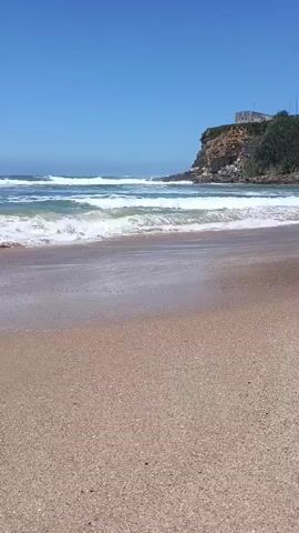 The video shows a view of a sandy beach with gentle waves rolling in. The sky is clear and blue. In the background, there is a rocky cliff with some vegetation and a building on top. The camera is positioned low to the ground, giving a sense of being right on the beach. The scene is peaceful and idyllic.