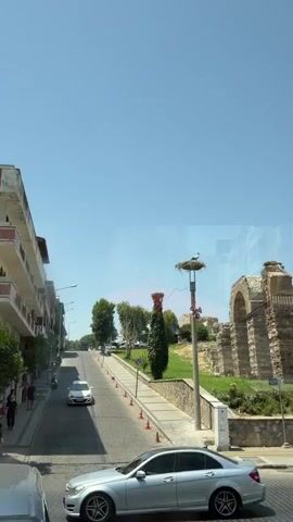 The video pans up a street on a clear, sunny day. A white car drives down the street, followed by a motorcycle with two passengers. On the right side of the street, there are ancient ruins and a tall lamppost with a stork's nest at the top. A stork is standing in the nest. Another, smaller stork's nest is visible behind the lamppost, on top of a conical bush. Buildings line the left side of the street.