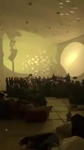 A well-lit, spacious room accommodates a large group of people. Some individuals are seated on the floor, utilizing pillows for comfort, while others are standing. In the front of the room, an orchestra and choir are performing. Projectors cast a bright yellow-green light onto the walls. One side of the room features a projection of small circles, while the other displays a larger, solitary circle. A speaker on a stand is positioned in the left corner of the room. The audience is focused on the orchestra.