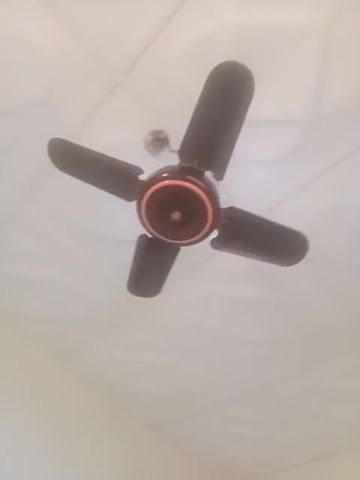 The video shows a ceiling fan with five dark brown blades and a central housing unit that is dark brown with an orange ring. The fan is mounted on a white ceiling. The blades are visible from below, and the fan appears to be stationary at the beginning of the clip, then begins to rotate slowly. The image quality is low, with some blur and graininess, making it difficult to discern fine details.