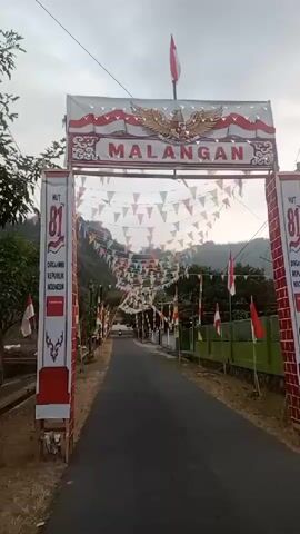 The video shows an archway decorated with Indonesian flags and banners. The archway is located at the entrance to a road lined with trees and houses. The road is paved and there are flags and pennants strung across it. The archway has the word "MALANGAN" written on it, along with an eagle emblem. On the sides of the archway, there are banners that say "HUT 81 DIRGAHAYU REPUBLIK INDONESIA". The sky is overcast.
