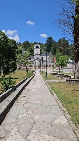 The video pans across a courtyard towards a large, stone religious building with a tall bell tower. A wide, paved pathway leads from the foreground to the building. The pathway is bordered on the left by a raised stone wall and grass. On the right, the pathway is bordered by grass, with several green lampposts spaced along it. In the background, the religious building is surrounded by trees and a rocky hillside. The sky is blue with a few white clouds. The sun is shining, indicating it is daytime. There are no people visible in the video.