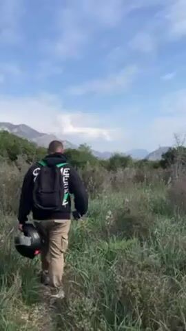 A man is walking through a grassy field toward a mountainous landscape under a blue and partly cloudy sky. He is wearing a black hoodie, tan cargo pants, and white sneakers. He has a black backpack on and is carrying a black motorcycle helmet in his left hand. The terrain is a mix of grass and low bushes, with some trees visible in the background and mountain peaks in the distance. The man is walking away from the camera.