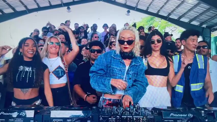The scene captures a vibrant outdoor party with a crowd of diverse individuals gathered under a large, roofed structure, possibly a pavilion or open-air venue. At the forefront, a DJ, a blonde woman with sunglasses and headphones around her neck, is actively performing at a Pioneer DJ console, seemingly mixing music. The crowd behind her is dancing, some holding beverages, indicating a relaxed and festive atmosphere. The attire of the partygoers varies, ranging from casual to more fashionable, with many sporting sunglasses. The setting suggests a sunny day with natural light illuminating the scene, and the greenery visible in the background hints at an outdoor location.
