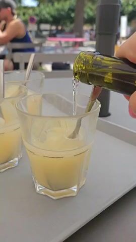 A hand holds a green glass bottle with a yellow-green liquid, pouring it into a clear faceted glass with a silver spoon inside. The glass is filled with a pale yellow liquid and ice cubes. Another glass with a similar drink is visible to the left. The glasses are on a gray tray. In the background, a person wearing a black baseball cap and a blue tank top is sitting at a table outdoors. Trees and possibly a building are visible in the distance. The lighting suggests it is daytime.