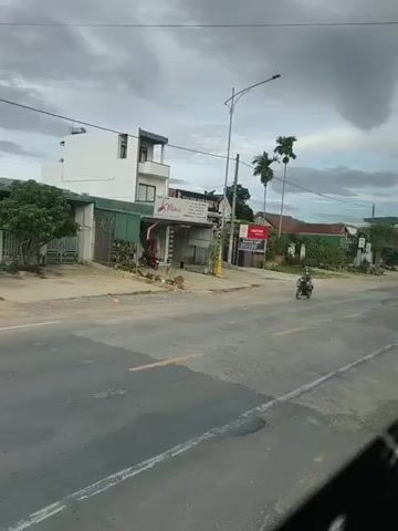 The video shows a road in what appears to be a rural or suburban area. The sky is cloudy. On the left side of the road, there are several buildings, including a white multi-story building, a smaller green building, and a more industrial-looking structure with an open front. There is also a tree and a fence. On the right side of the road, there are more buildings and what looks like a small business, possibly a motorcycle repair shop, with several motorcycles parked outside. A person wearing a blue and white patterned shirt and a face mask is riding a black motorcycle in the foreground, moving from left to right across the screen. Another person is seated on the ground in front of the business on the right. The road surface is asphalt with yellow lane markings.
