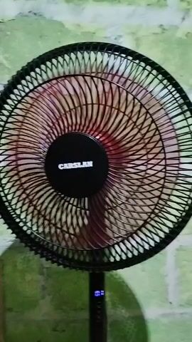 A close-up shot shows a black standing fan with a red center and the brand name "CARSLAN" in white lettering. The fan's blades are blurred due to their rapid rotation, indicating that the fan is on. A digital display on the fan's stand shows "01" in blue light. The fan is positioned in front of a textured, greenish-white wall. A shadow of the fan is cast on the wall. The lighting is somewhat dim, and the video is shaky.