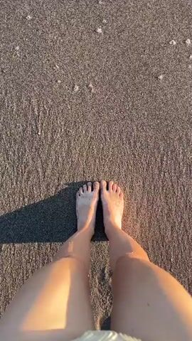 The video shows a pair of bare feet standing on a beach. The sand is dark and wet, with small waves washing over the feet. The perspective is from above, looking down at the feet and the encroaching water. The light suggests it is either early morning or late afternoon, casting a shadow to the left of the feet. A portion of a patterned bikini bottom is visible at the bottom of the frame. The water is clear and foamy as it recedes.