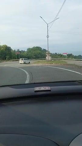 The video shows a view from the dashboard of a car as it drives on a winding road. The road has white lane markings and is bordered by trees and foliage. There are several red and white chevron signs indicating sharp turns in the road. A white van is visible parked on the side of the road in the distance. The sky is overcast with some clouds. On the dashboard, there are two air fresheners and a car charger. The car appears to be moving at a moderate speed.