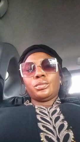 A woman is sitting in a car and recording herself. She is wearing sunglasses and a headscarf. The car interior is visible in the background.