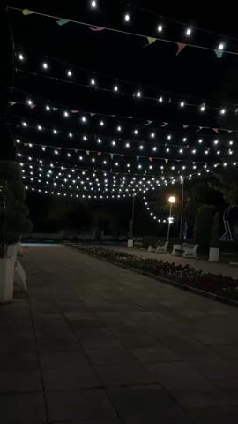 The video shows a serene night scene in a park or pedestrian area in Smirnenski, Bulgaria. The setting is illuminated by numerous string lights strung across the pathway, creating a festive canopy effect. Small, colorful triangular flags are interspersed with the lights. The pathway is paved with dark-colored square tiles.

Along the sides of the path, there are long flowerbeds filled with red and white flowers. Park benches are visible at various points, suggesting places for relaxation. Several lampposts cast a warm glow, further lighting the area. In the background, there are trees and shrubs, silhouetted against the night sky. The overall atmosphere is peaceful and inviting, likely captured on a clear night. No people or specific activities are visible, emphasizing the quiet ambiance of the park after dark. There is no visible text.