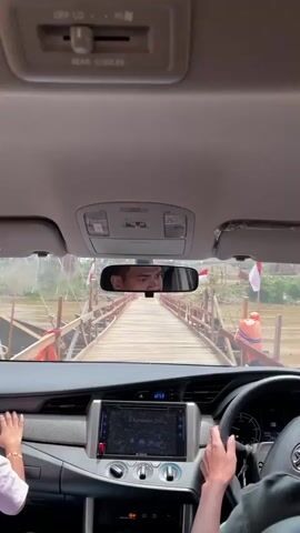 The video is filmed from inside a car, looking forward through the windshield and at the interior dashboard. The car is approaching and driving onto a wooden bridge that spans over water. The bridge has pink railings and appears to be a tourist attraction, with Indonesian flags visible on either side. The dashboard of the car shows a navigation screen and various controls. In the driver's seat, a person's hands are visible on the steering wheel. In the passenger seat, a child's feet are visible. The rearview mirror shows the face of a person, presumably the driver. The scene is bright and sunny.