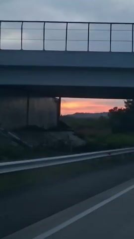 The video shows a view from a moving vehicle on a highway. The camera is focused on the sky and the landscape seen through an opening, possibly under an overpass. The sun is setting, casting a warm orange and pink glow across the sky, with some clouds present. Trees and vegetation are visible on the right side of the frame, silhouetted against the colorful sky. A guardrail runs along the side of the highway. The perspective suggests the vehicle is traveling forward, and the overpass is passing overhead.