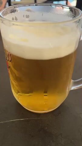 A close-up shot shows a glass mug filled with beer and a thick layer of foam. A person's finger is visible in the foreground, touching the side of the mug. The background is out of focus but appears to be an outdoor setting with some people and structures. The lighting is natural, suggesting it might be daytime.
