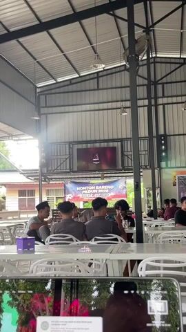 A group of young men are seated at white tables in an open-air, covered dining area. They appear to be watching a large screen displaying a sports event, possibly football, as suggested by the banner behind them that reads "NONTON BARENG PILDUN 2026 BOLA GEMBIRA." Sunlight streams in from the left side of the frame, illuminating dust motes in the air and creating lens flare. The men are casually dressed, some are looking at the screen, while others are interacting amongst themselves. One man in the foreground is seen holding a phone or camera, with its screen displaying a colorful image. The overall atmosphere seems relaxed and social.