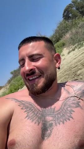A man is lying on a beach and laughing while looking up at the sky. The man has dark hair and a beard, and he is wearing a gold chain with a cross pendant. He has a large tattoo of a skull with wings on his chest. The beach is sandy and there is a hill with green vegetation in the background. The sky is blue.