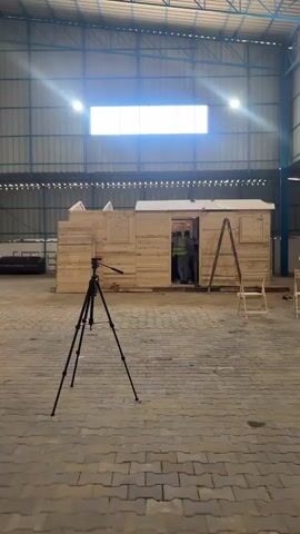 The video shows a construction site inside a large, open-air warehouse or industrial building. A wooden structure, resembling a small house or modular unit, is under construction in the center of the frame. Several people, some wearing safety vests, are working on and around the wooden structure, suggesting an ongoing building or assembly process. A tripod with a camera is positioned in the foreground, indicating that the scene is being filmed or photographed. A ladder leans against the wooden structure, and a simple wooden chair is placed nearby. The building has a corrugated metal roof and a large, illuminated window or skylight at the top, suggesting it is daytime. The floor is paved with interlocking bricks. The overall atmosphere appears to be one of active construction and work. There is no visible text in the media file.