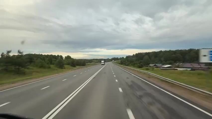 The video shows a view from the driver's seat of a vehicle traveling on a two-lane highway. The highway is paved and has white lane markings. On either side of the highway are grassy shoulders with guardrails. Beyond the shoulders are trees and some rural buildings. The sky is cloudy with patches of light suggesting either sunrise or sunset. There are two trucks visible on the highway. One truck, a white box truck, is moving towards the camera in the left lane. The second truck, a white tractor-trailer, is moving away from the camera in the right lane, further in the distance. The terrain appears to be gently rolling hills.