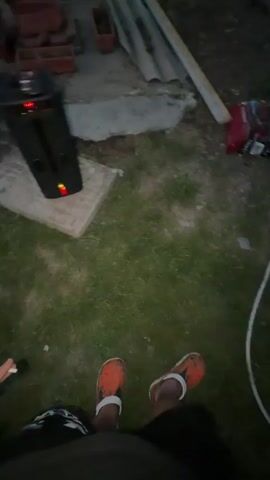 The video shows a person from the knees down, wearing orange Crocs, standing in a grassy area at dusk. In the background, a charcoal grill is lit and ready for cooking. A raw cut of meat, a bottle of beer, and other items are on a side table next to the grill.