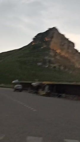 The video shows a mountain with lights illuminating its side. In the foreground, there are two people walking away from the camera, and a road. There are also some stalls or small buildings on the side of the road. The sky is cloudy, and it appears to be dusk or dawn. A car is seen driving on the road.