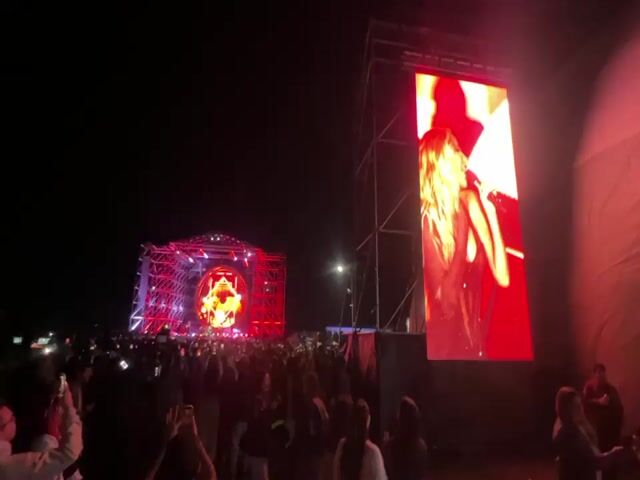 The video shows an outdoor music concert at night. A large crowd of people is gathered in front of a stage, many holding up their phones to record. The stage is lit up with red lights and features a circular design in the center. To the right of the stage, there is a large screen displaying a live feed of a woman singing on stage. The screen occasionally switches to show a band member playing the guitar. The overall atmosphere is energetic and lively.