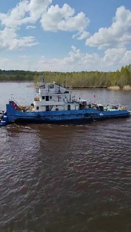 The video shows a blue and white tugboat traveling on a river under a partly cloudy sky. The tugboat has a Russian flag on the back. It is pushing a raft of logs. The riverbanks are lined with trees.
