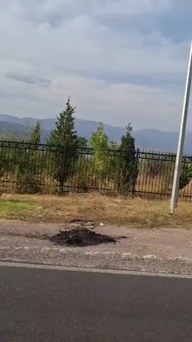 The video captures a panoramic view of a landscape from a moving vehicle. In the foreground, a paved road runs horizontally across the bottom of the frame. Beyond the road, a dirt shoulder leads to a black metal fence. Behind the fence, sparse vegetation and trees are visible. In the mid-ground, a body of water is partially obscured by the fence and foliage. The background features rolling hills and mountains under a cloudy sky. The camera pans from left to right, showing the continuous scenery. There are no people or animals visible in the video.