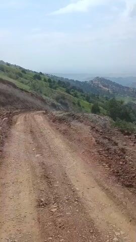 The video shows a view from a moving vehicle on a dirt road. The road is rough and appears to be undergoing some kind of construction or repair, with piles of dirt and rocks on the side. The vehicle is driving uphill, and the landscape is mountainous with green hills and scattered trees. The sky is hazy and overcast. The camera is looking forward and slightly to the right. The video appears to be taken during the day.