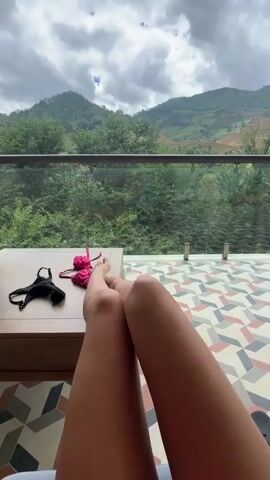 A person is relaxing on a balcony with a scenic mountain view. The camera pans from the person's legs and feet, where two bikinis are resting on a wooden table, to reveal a breathtaking landscape of lush green mountains under a partly cloudy sky. The balcony has a glass railing and a tiled floor with a geometric pattern.