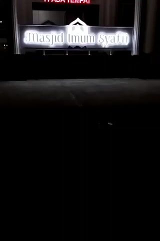 A brightly lit sign reading "MASJID UMUM SYADIL" is prominently displayed at the entrance of a mosque at night. The sign is white with black lettering and illuminated with white light. Above the sign, another red neon sign reads "PARKIRLAH". The mosque facade is white with arched doorways and Islamic calligraphy. The surrounding area is dark, suggesting it is nighttime.