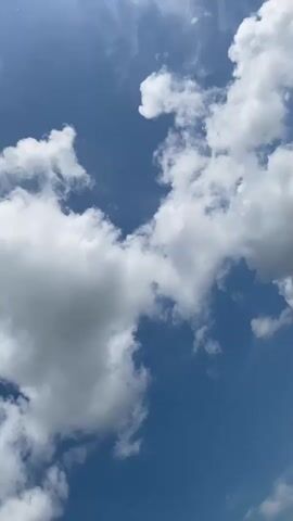 The media file is a short, vertical video of a blue sky with white, fluffy clouds. The camera is pointed upwards, capturing a dynamic display of cloud formations. Some clouds are large and dense, while others are wispy and scattered. The lighting suggests it is daytime, with bright sunlight filtering through the clouds, creating varying degrees of shadow and brightness. There is no discernible activity or event taking place; the focus is purely on the natural beauty of the sky. The weather appears to be fair with some cloud cover. The provided location context indicates this is likely filmed in Grigoryevskoye, Russia. No people, objects, or text are visible in the video. The overall emotion evoked is one of peacefulness and expansiveness, common when observing the sky.
