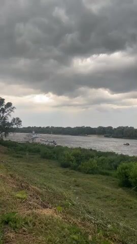 The video shows a wide river under a cloudy sky. On the left bank, there is a structure with boats docked nearby, and beyond that, a line of trees. The right bank of the river is also lined with trees, forming a dense forest. In the foreground, there is a grassy slope with some dry brush. There are a few small boats visible on the river. The overall impression is of a natural landscape with some human activity.