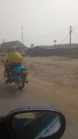 The video shows a view from inside a car driving on a paved road through a dusty, somewhat rural area.  There's a building on the left with a long, covered porch area, and piles of what looks like refuse or construction materials in front of it.  A white truck is parked nearby.  A tall cell tower is visible in the background.  A motorcycle passes the car on the left, and a man walks in the opposite direction on the side of the road.  Later, there's a small palm tree in the distance. Several people are walking around in the background. The air seems hazy.