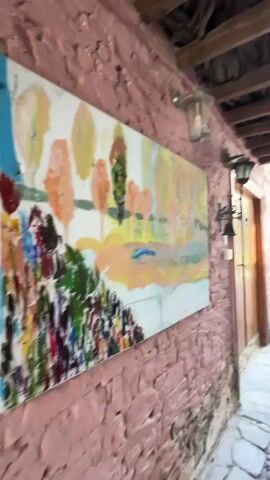 The video pans to the right, showing a narrow cobblestone alleyway. The walls on either side are made of stone and painted in muted colors. A large painting of a landscape hangs on the left wall. On the right wall, a display of books is set up, and a potted plant sits on a small table. Vines grow along the wall, adding a touch of greenery. Sunlight streams into the alley from the opening at the end, illuminating the scene.