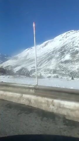 The video shows a snowy mountain landscape under a clear blue sky, as seen from a moving vehicle. The mountain is covered in snow and sparse vegetation. There is a road with a guardrail visible in the foreground. The camera appears to be inside the vehicle, looking out at the scenery.  The video is shaky due to the movement of the vehicle. The overall impression is a cold, wintery scene.