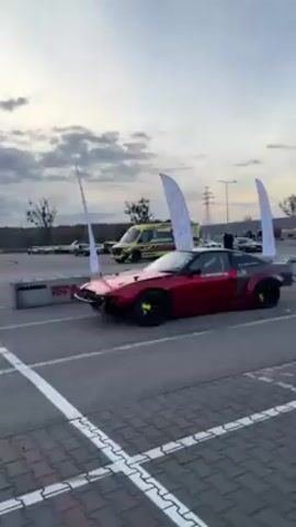 The video shows a red and gray drift car idling in a parking lot with other cars in the background. The drift car has a large spoiler and neon green accents, and is parked near a tire barrier and some white flags. The parking lot is marked with white lines, and the weather is overcast.