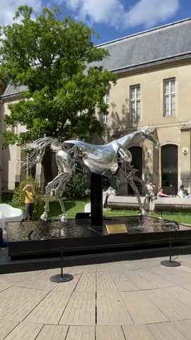 A life-sized, metallic horse sculpture is displayed outdoors in a courtyard. The sculpture is detailed and appears to be made of polished metal, reflecting the surroundings. It is positioned on a black base in a shallow pool of water. In the background, a building with large windows and a grey roof is visible. Several people are present in the scene. To the left of the sculpture, a person in a yellow shirt stands near the water. Further back, a few people are seated and interacting. To the right of the sculpture, two people are standing. Lush green trees and a cloudy sky complete the outdoor setting. The ground in front of the sculpture has a circular pattern with metal grates.