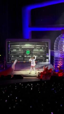 The video shows a concert performance on a stage. A female singer is center stage, dressed in a white outfit and holding a microphone. Behind her, a large screen displays a graphic that resembles a computer control panel with many buttons and indicators. To the singer's right, a giant, clear plastic water bottle with a blue label stands prominently. In the foreground and to the right of the singer, several performers in red outfits are positioned on the stage. The audience is visible in the dark foreground, with many holding up their phones, suggesting they are recording or photographing the event. The stage is lit with blue and purple lighting, creating a vibrant atmosphere.