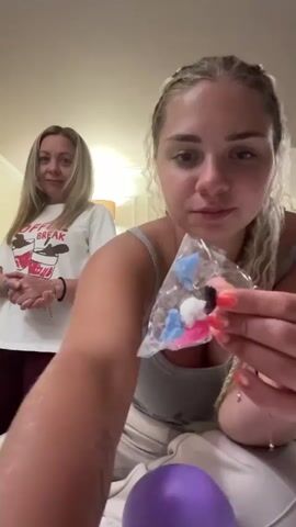 A young woman with blonde braided hair, wearing a grey tank top and adorned with rings and bracelets, holds a small plastic bag containing colorful, small objects, possibly accessories for a balloon. Her fingernails are painted neon orange. Her hands are in focus as she manipulates the bag. Behind her, to the left, an older woman with blonde hair, wearing a white t-shirt with a graphic print and dark leggings, stands with her arms crossed. She is looking towards the camera with a slight smile. The scene appears to be indoors, possibly a bedroom, with soft lighting and neutral-colored furniture. A large purple object, likely a deflated balloon, is visible at the bottom center of the frame.