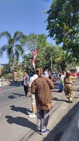 The video shows a street parade, likely celebrating Indonesia's Independence Day. People are walking down a paved street lined with palm trees and other foliage, under a clear blue sky. On the left, a man in a brown and black striped shirt and light gray pants is facing away from the camera. A person in a military-style uniform walks parallel to the parade, gesturing, and holding a two-way radio. At least one other person is waving an Indonesian flag. On a truck, someone stands on top of large speakers, dancing, and waving their arms. A banner on the truck reads "Dirgahayu Indonesia" and "17 Agustus 1945 - 17 Agustus 2023". People dressed in traditional Indonesian clothing are part of the parade, and a few onlookers are seated on the curb on the right. The sounds of music and celebration are present in the background.